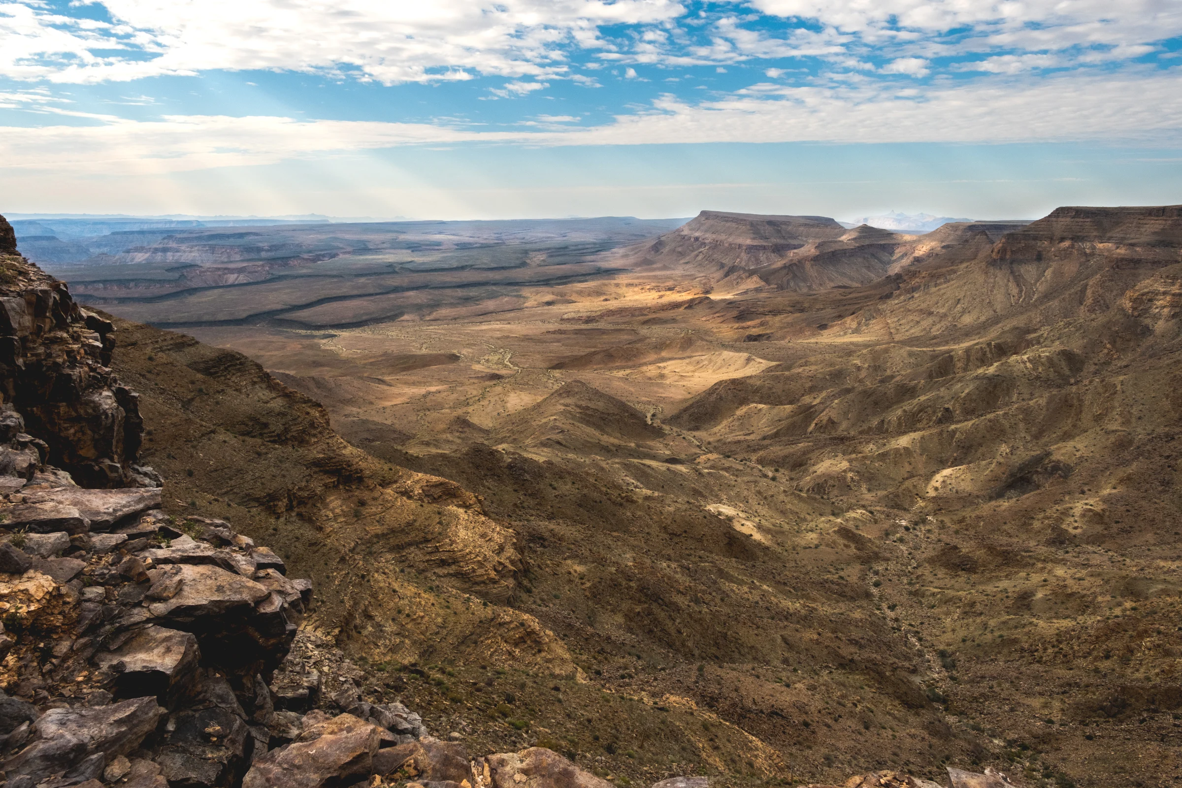 魚河峽谷 Fish River Canyon|層疊崖壁、谷地與遠方台地展開,雲隙間的光勾勒地形深度。